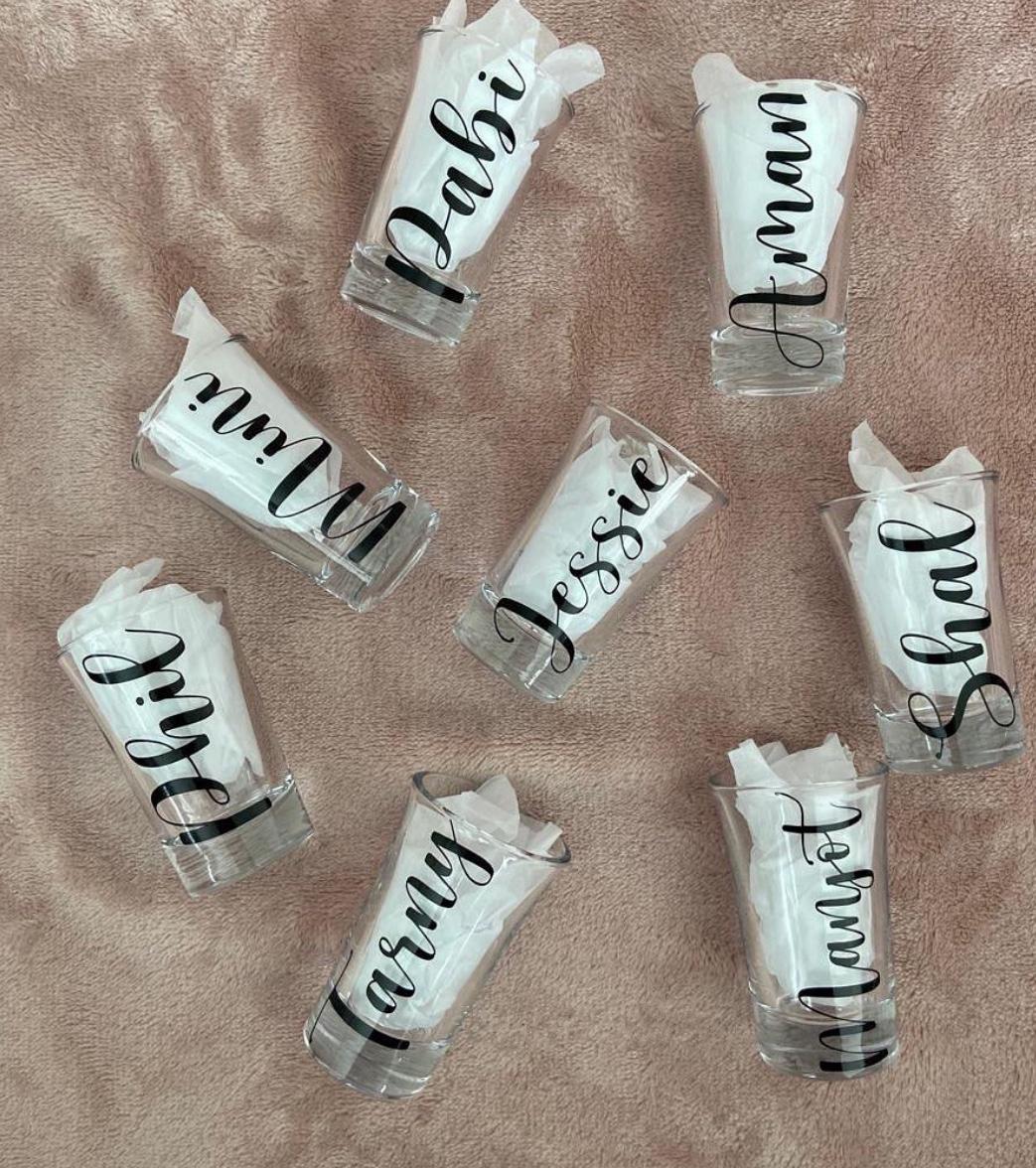 Multiple custom shot glasses with black calligraphy name vinyl, arranged on a blush velvet surface